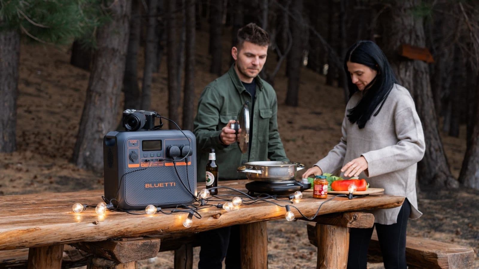 A Bluetti portable power station on a wooden table at a campsite, powering string lights and a hob while two people prepare a meal, with a camera resting on top of the unit.