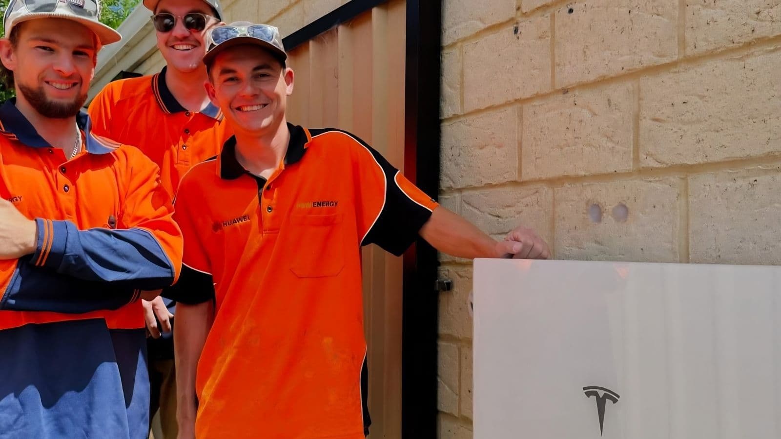 Three installers in high-visibility polos standing beside a newly installed wall-mounted Tesla Powerwall